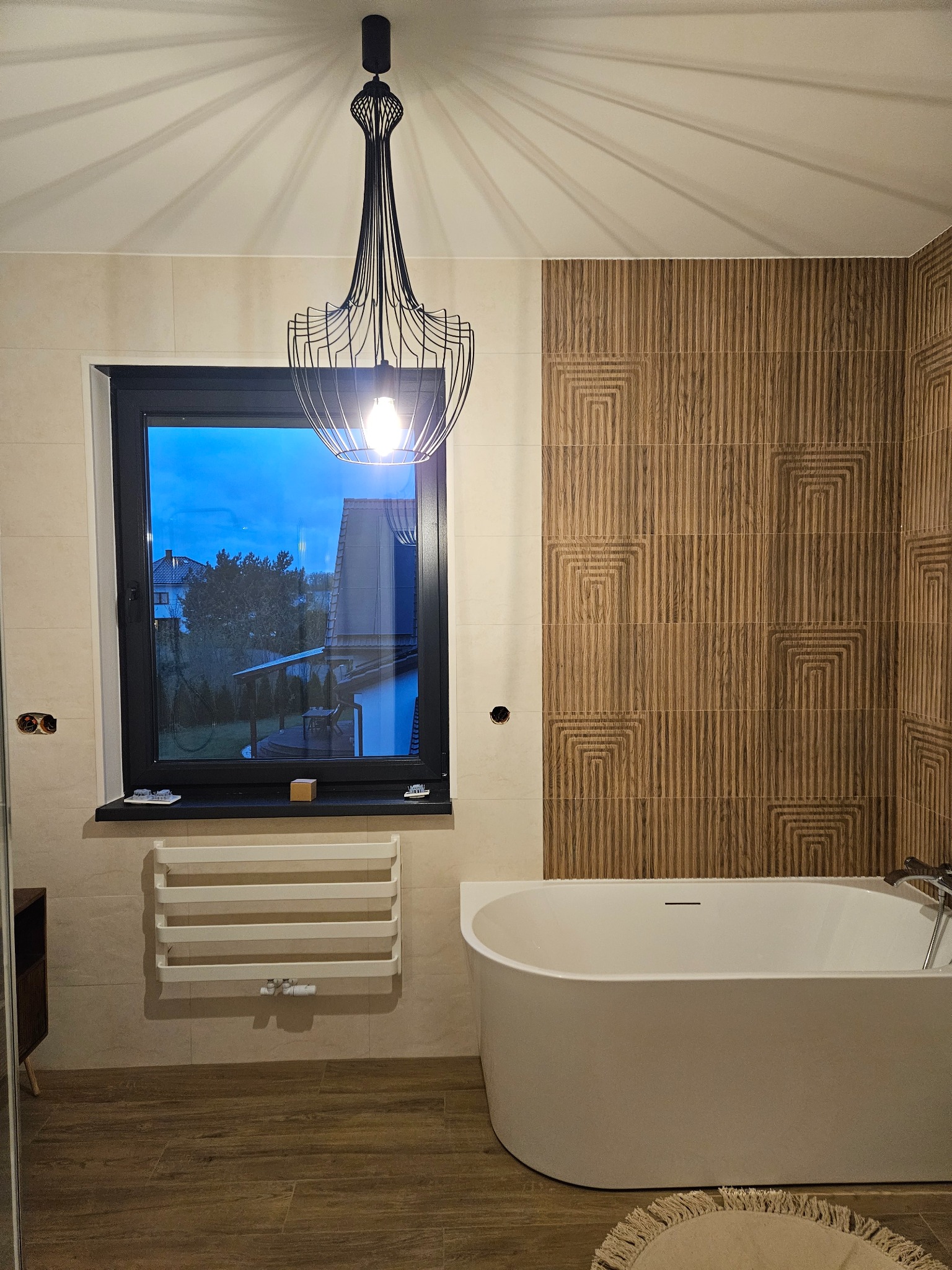 Nowoczesna łazienka z wolnostojącą wanną, drewnopodobnymi płytkami i designerską lampą wiszącą, widok na okno z wieczornym krajobrazem.