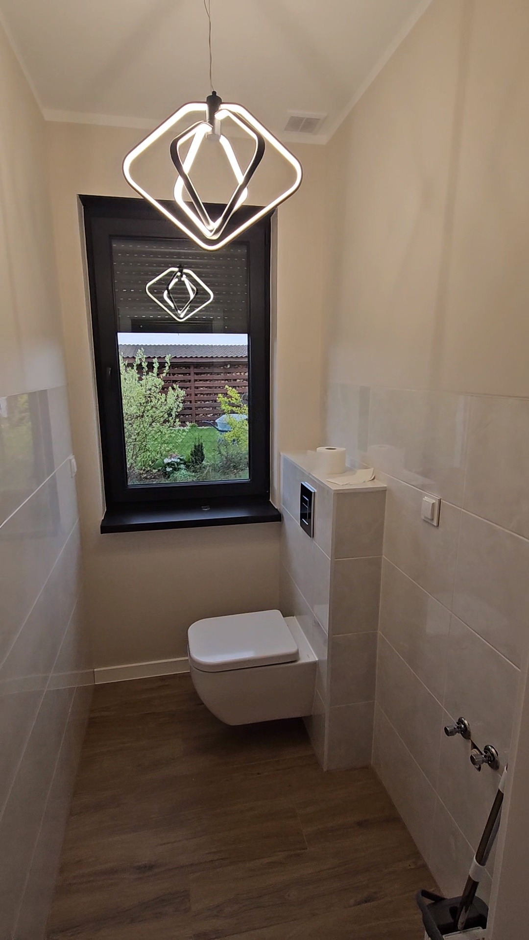 Nowoczesna łazienka z podwieszaną toaletą, jasnymi płytkami i oknem z widokiem na zieleń. Uwagę przyciąga designerska lampa sufitowa o geometrycznym kształcie.
