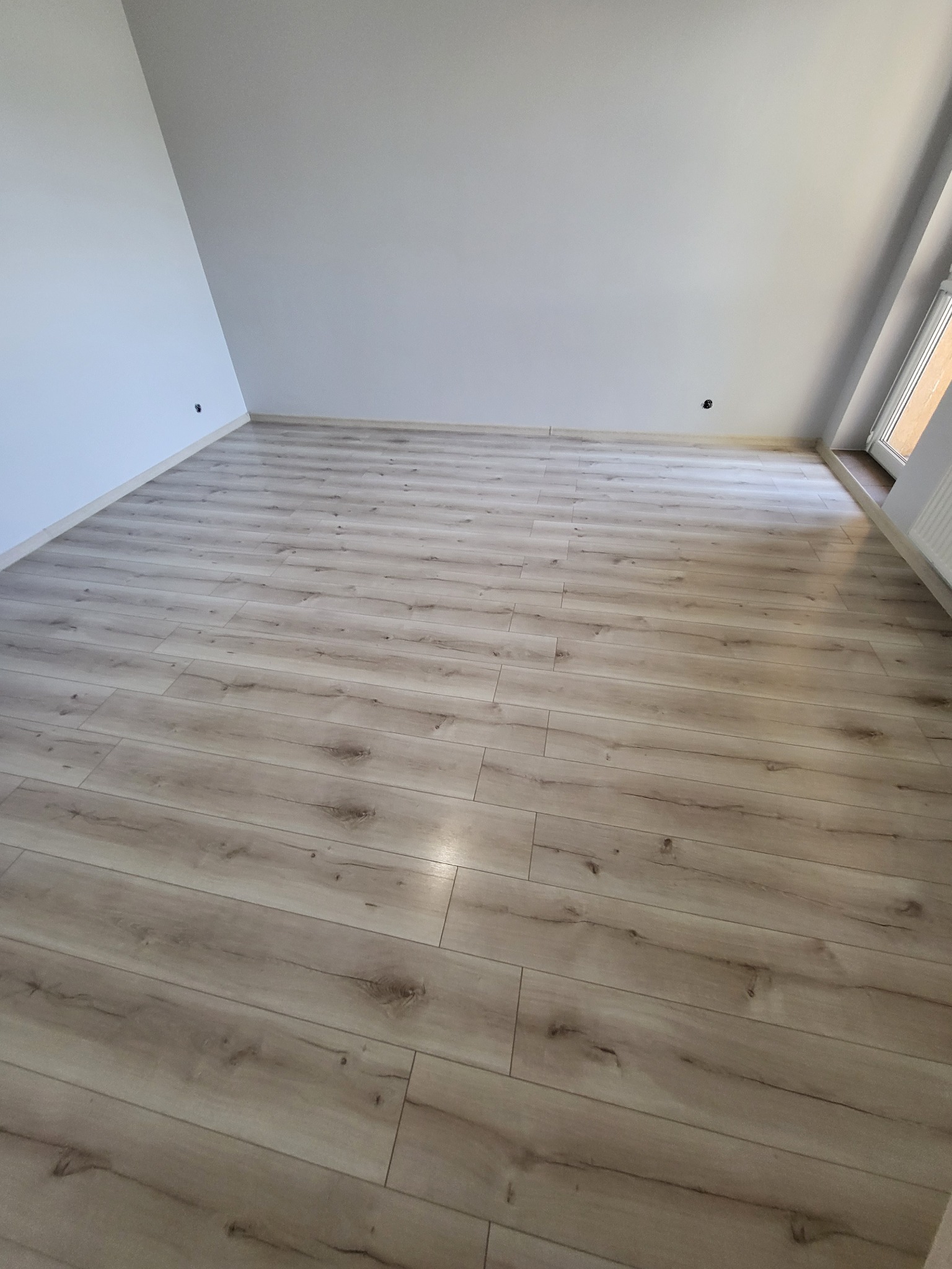 Jasne panele podłogowe ułożone w pokoju na poddaszu, widoczne gniazdka elektryczne przy podłodze, okno w prawym górnym rogu.