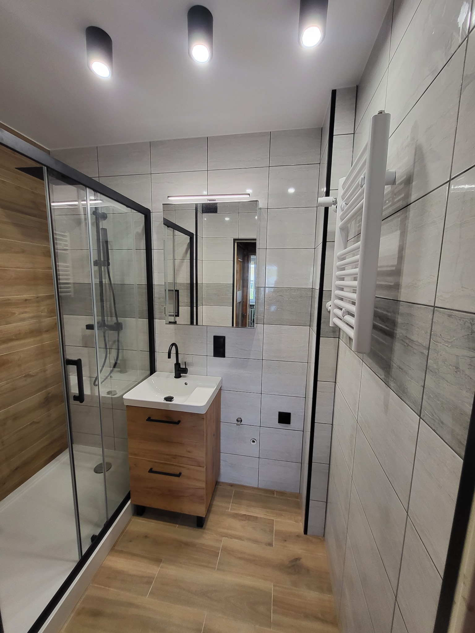Nowoczesna łazienka po remoncie: kabina prysznicowa z czarną ramą, drewniana szafka pod umywalką z czarną armaturą, grzejnik drabinkowy i płytki w odcieniach szarości i drewna.