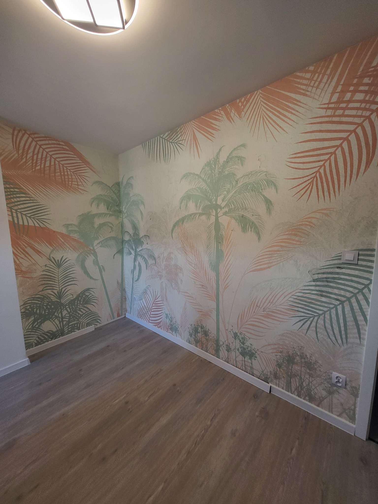 Pokój po tapetowaniu z motywem tropikalnych palm i drewnianą podłogą. Widoczne gniazdko elektryczne i listwy przypodłogowe. Oświetlenie sufitowe.