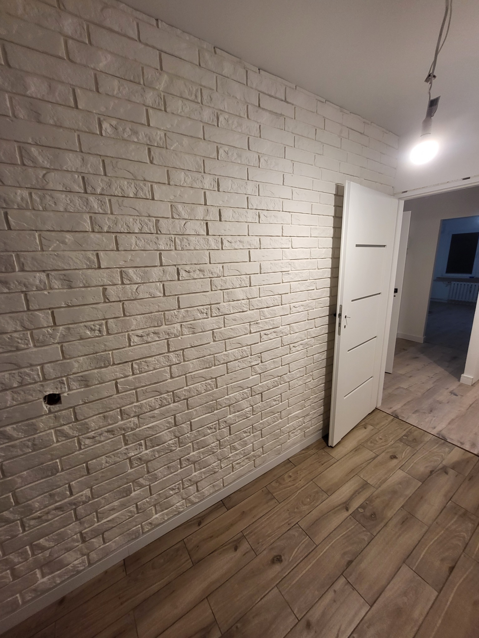 Ściana z białej cegły dekoracyjnej i drewnopodobna podłoga, widok na otwarte drzwi do kolejnego pomieszczenia. Widoczny fragment instalacji elektrycznej.