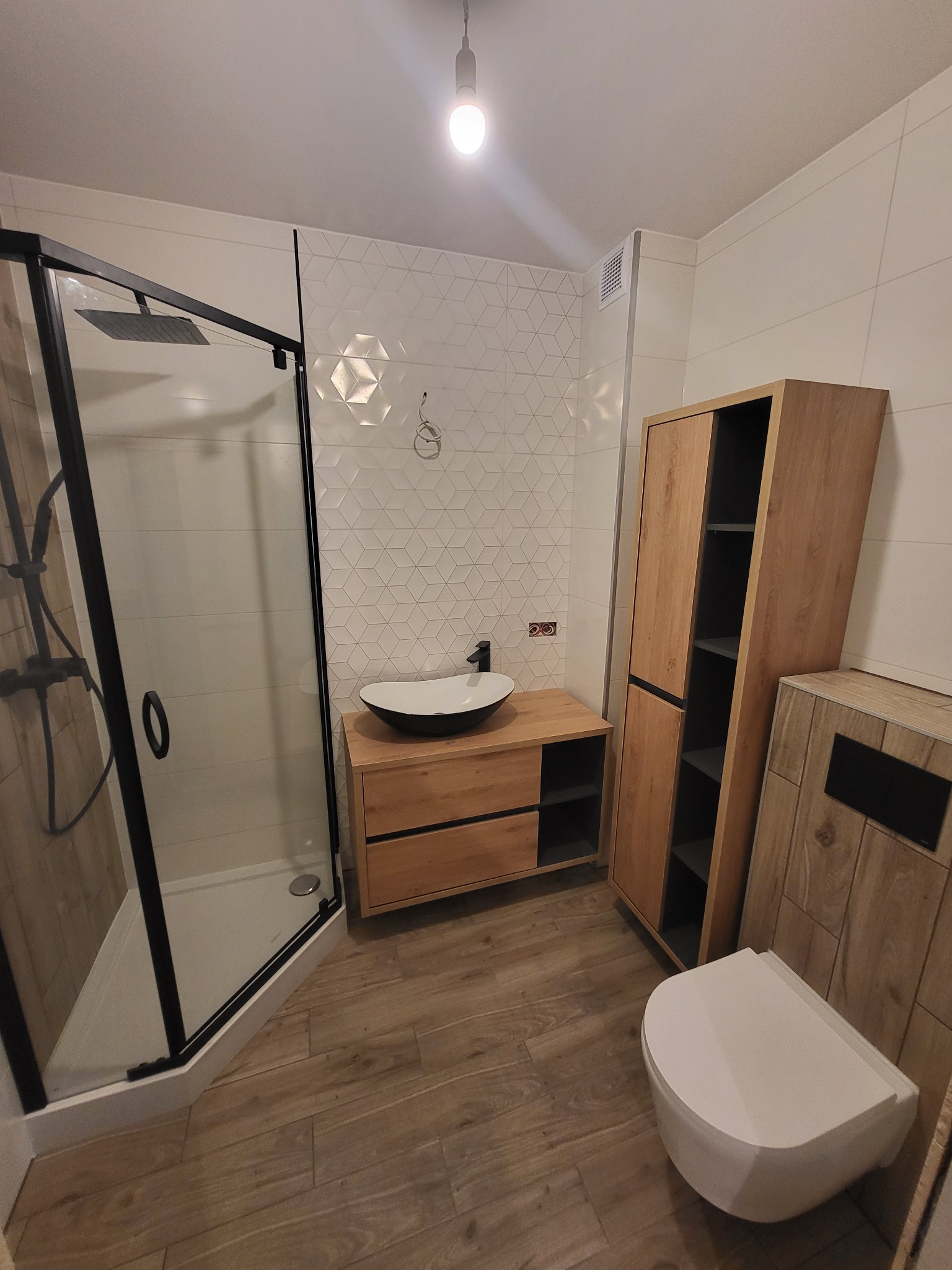 Nowoczesna łazienka z kabiną prysznicową typu walk-in, drewnianą szafką pod umywalką i wysoką szafką. Białe płytki heksagonalne na ścianie.