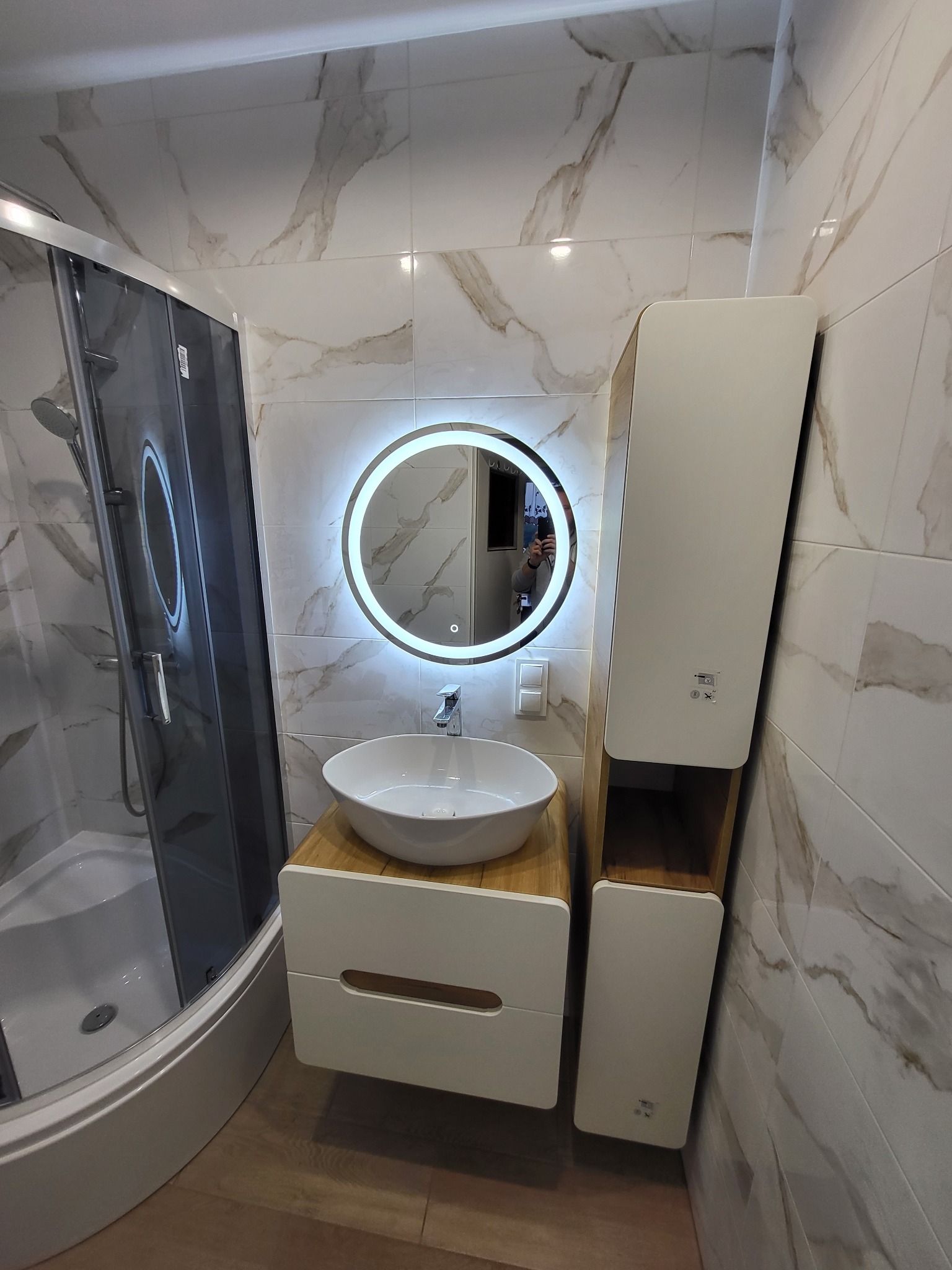 Nowoczesna łazienka po remoncie: kabina prysznicowa, umywalka z szafką z drewnianym blatem i podświetlane lustro na tle marmurowych płytek.