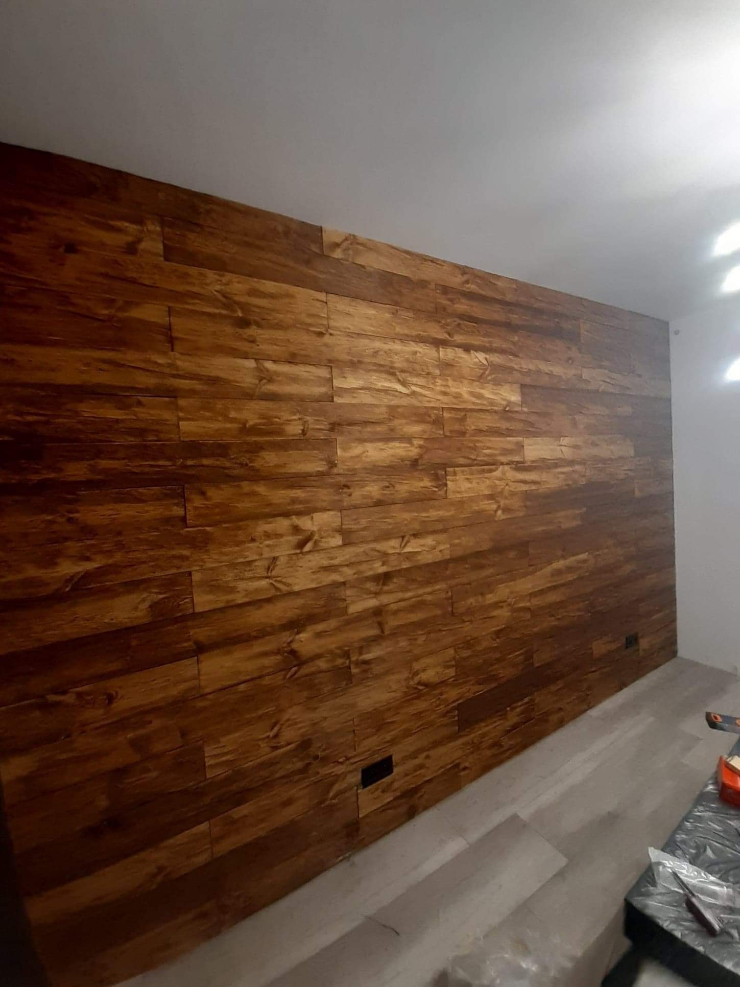 Ściana wykończona panelami drewnianymi w odcieniach brązu, ułożonymi poziomo. Widoczne fragmenty podłogi i sufitu. Nietypowy wzór paneli z wyraźnym usłojeniem.