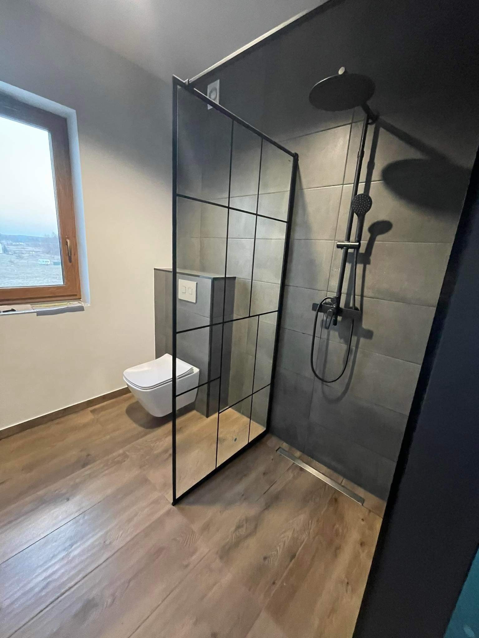 Nowoczesna łazienka z kabiną typu walk-in, szare płytki, drewnopodobna podłoga, czarna armatura i widok z okna na krajobraz miejski w tle.