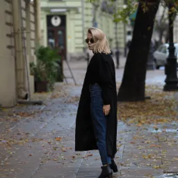 Kobieta w czarnym płaszczu i okularach przeciwsłonecznych spogląda przez ramię na tle jesiennego pejzażu miejskiego z opadłymi liśćmi na chodniku.