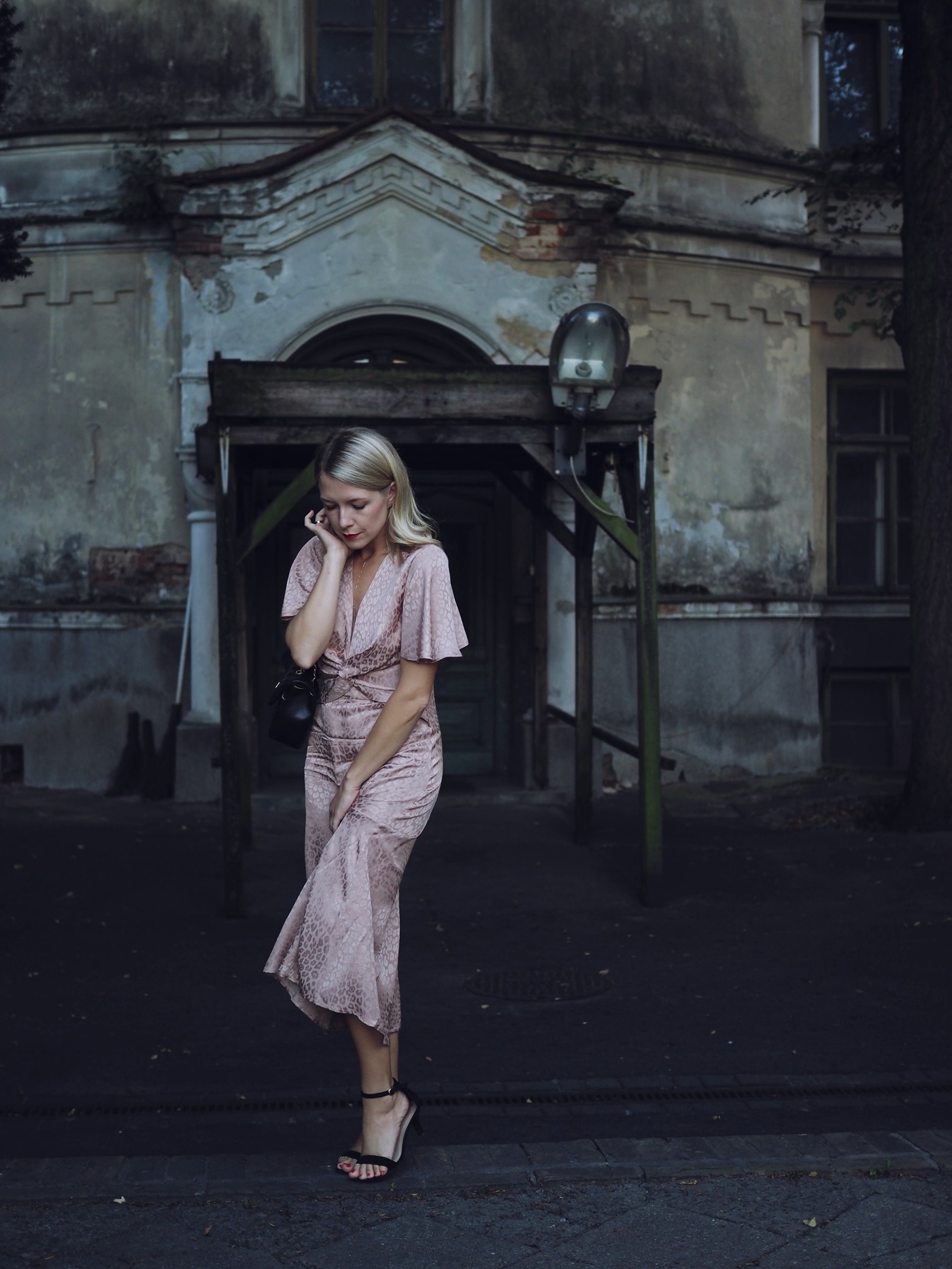 Blondwłosa kobieta w różowej sukience pozuje przed starą, zaniedbaną kamienicą z podświetlonym wejściem.