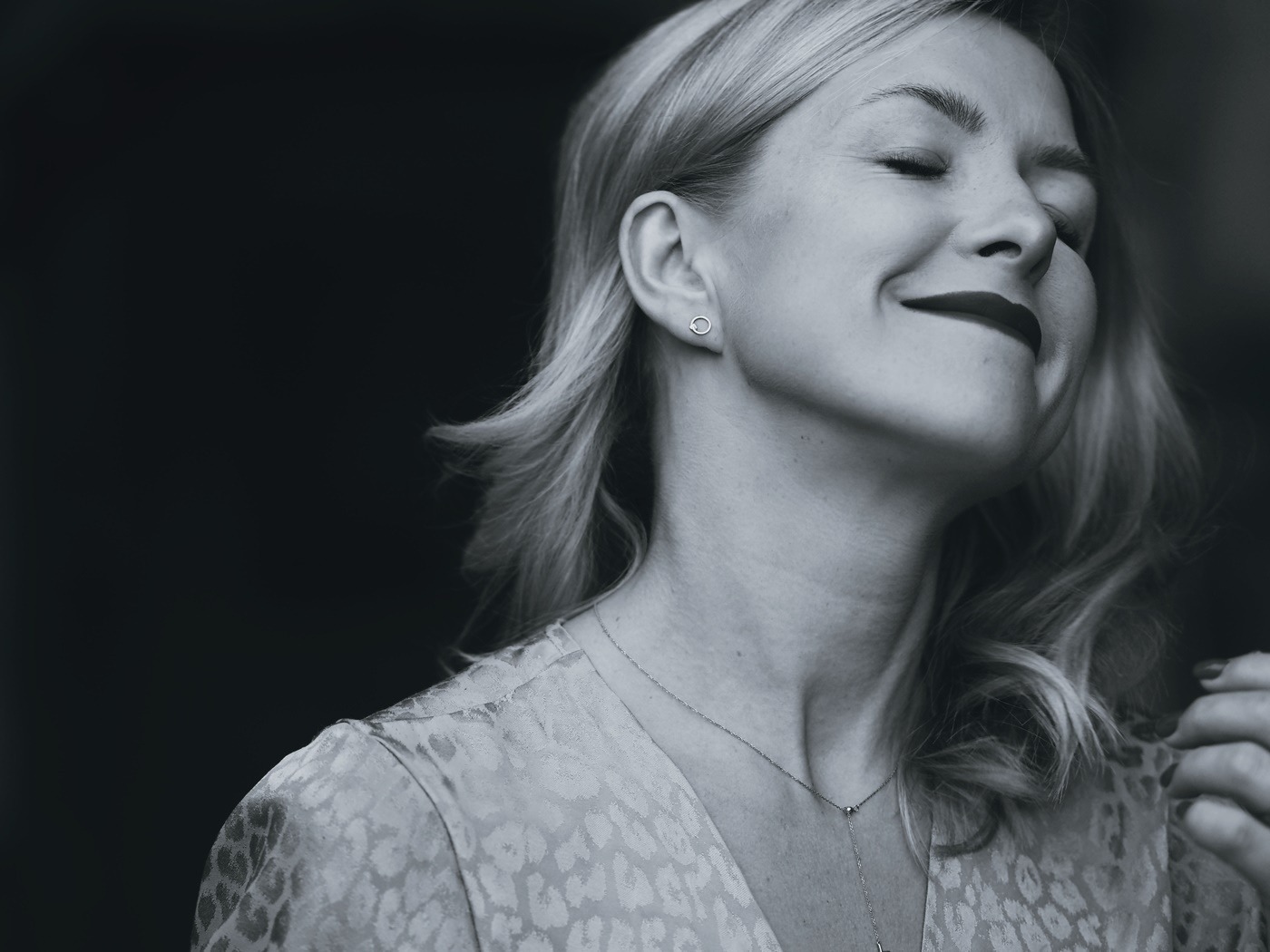 Portret kobiety z zamkniętymi oczami, subtelnym uśmiechem i delikatną biżuterią na szyi i w uchu, sfotografowanej w czerni i bieli z ciemnym tłem.