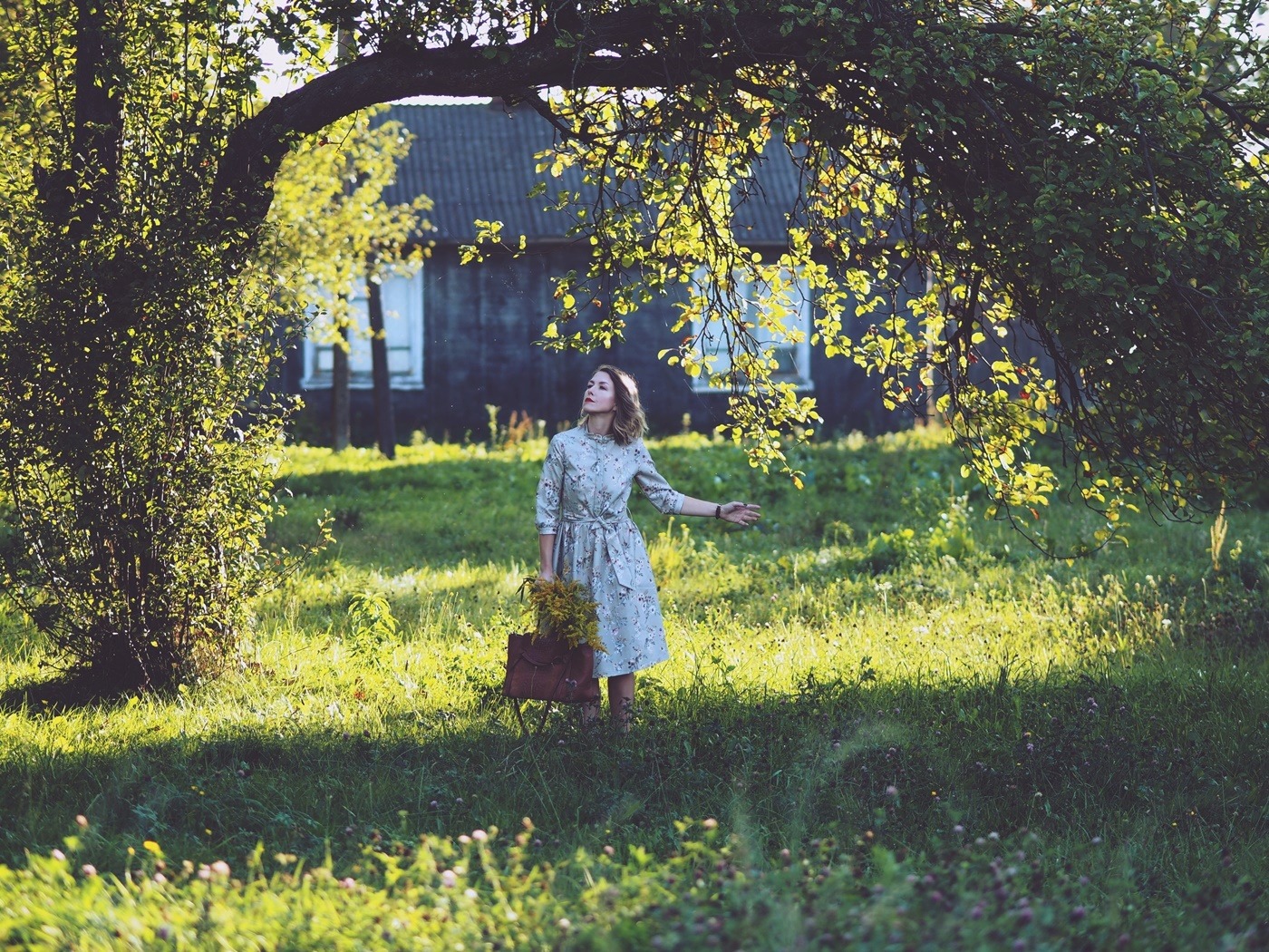 Kobieta w kwiecistej sukience stoi na łące pod drzewem, trzymając skórzaną torbę z polnymi kwiatami, w tle widoczny drewniany dom.