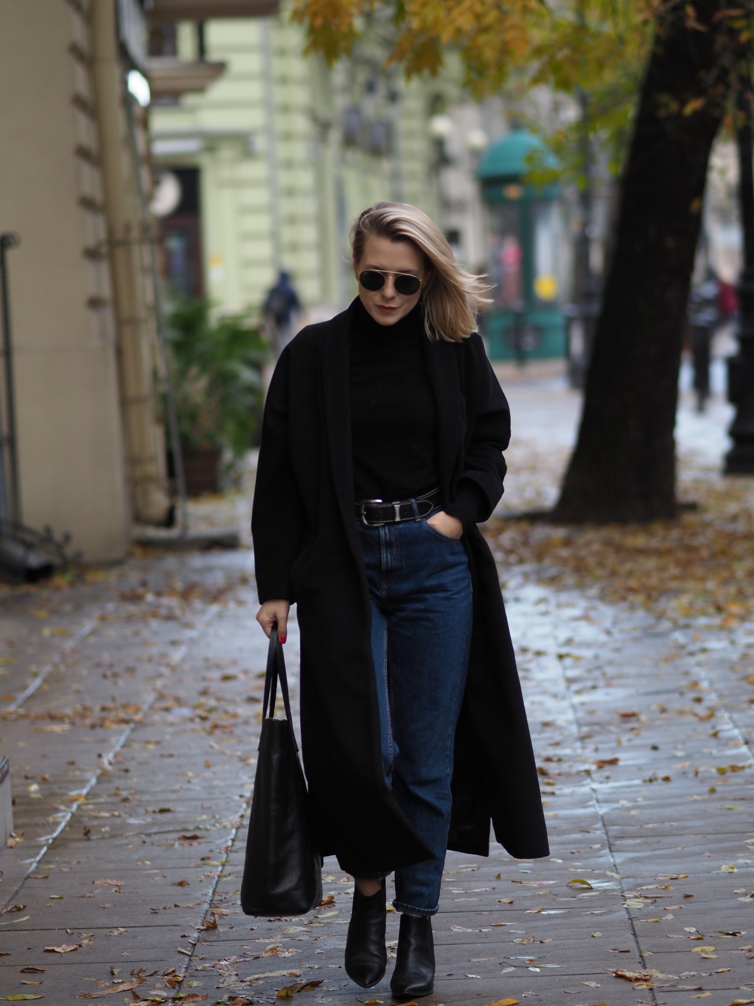 Kobieta w czarnym płaszczu i jeansach idzie chodnikiem usianym liśćmi, trzymając czarną torbę na tle miejskiej zabudowy i jesiennych drzew.