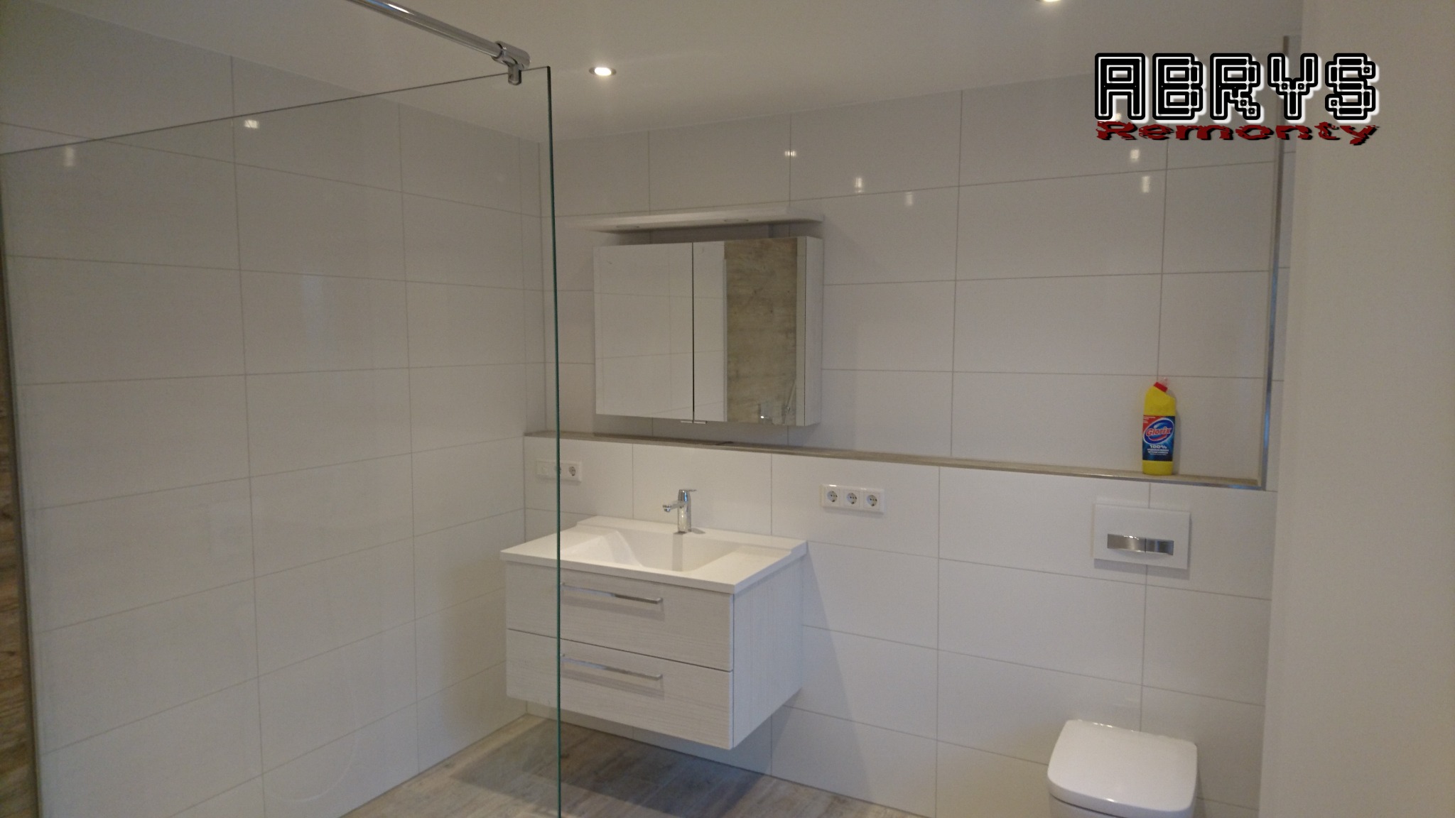 Nowoczesna łazienka z białymi kwadratowymi płytkami, podwieszaną szafką z umywalką, lustrem w szafce i żółtą butelką środka czyszczącego na półce; widoczne logo firmy w rogu.