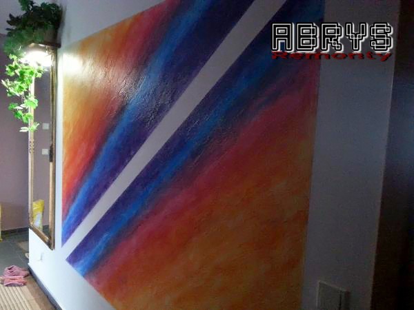 Wielobarwna ściana z abstrakcyjnym wzorem w odcieniach pomarańczy, czerwieni i niebieskiego, przecięta białym pasem, z logo firmy 'ABRYS Remonty' w prawym górnym rogu. W tle widoczna roślina...