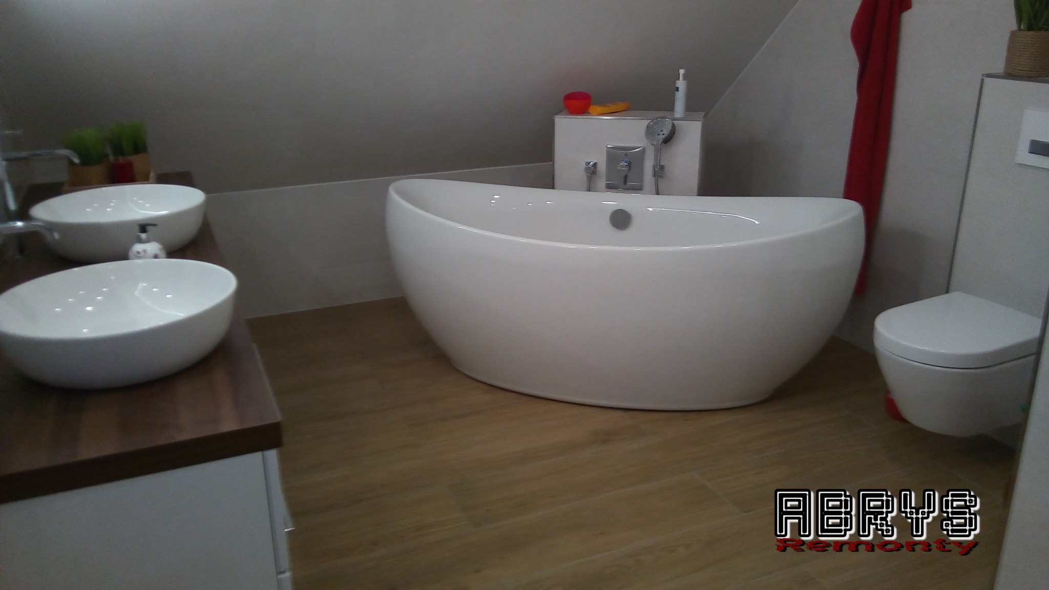 Nowoczesna łazienka z wolnostojącą wanną, dwiema umywalkami nablatowymi na drewnianym blacie i podwieszaną toaletą, wykończona w jasnych kolorach z drewnopodobną podłogą.