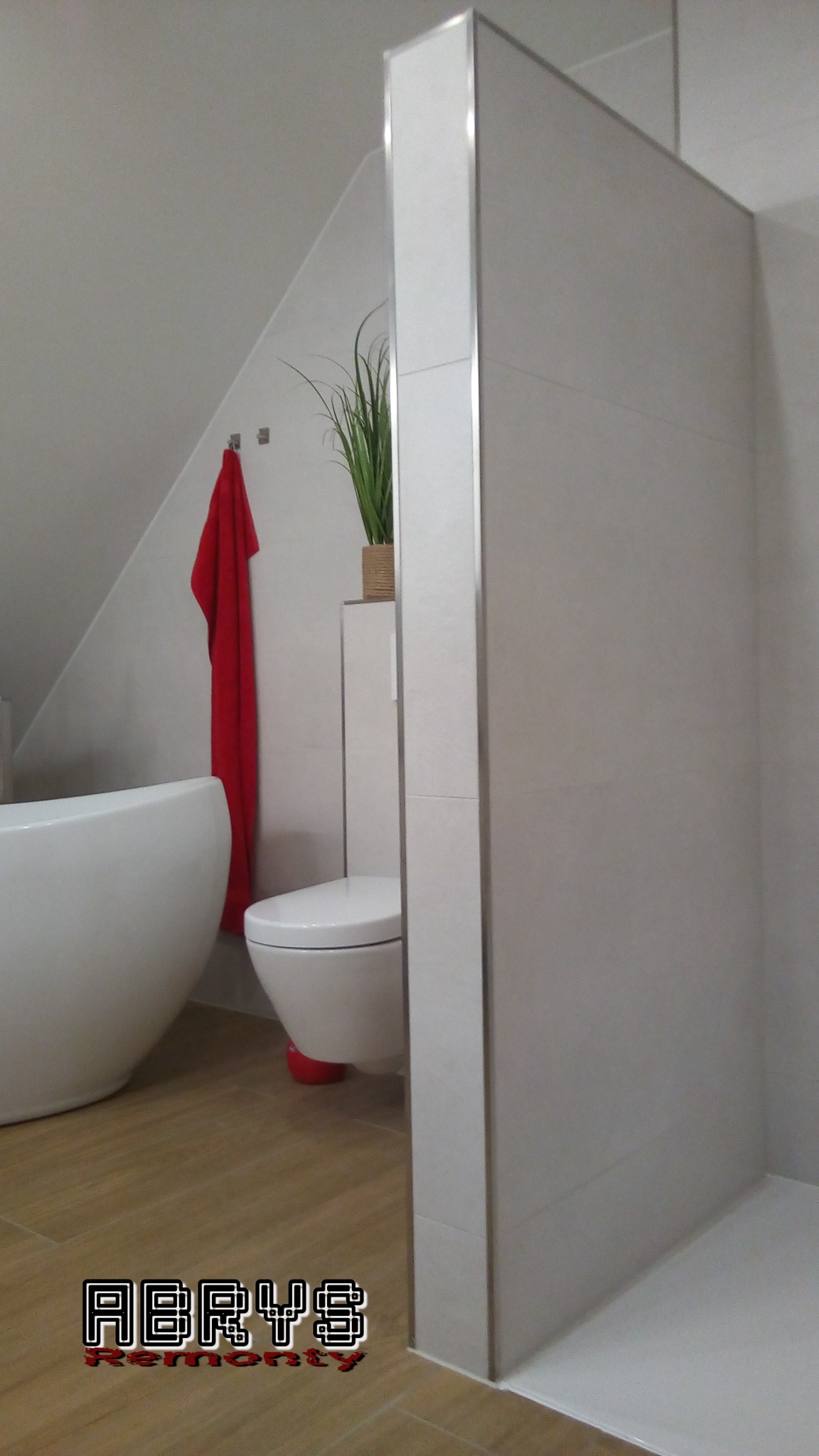 Nowoczesna łazienka z beżowymi płytkami, kabiną prysznicową typu walk-in, wiszącą toaletą z czerwonym elementem, wolnostojącą wanną i czerwonym ręcznikiem na wieszaku pod skosem dachu.