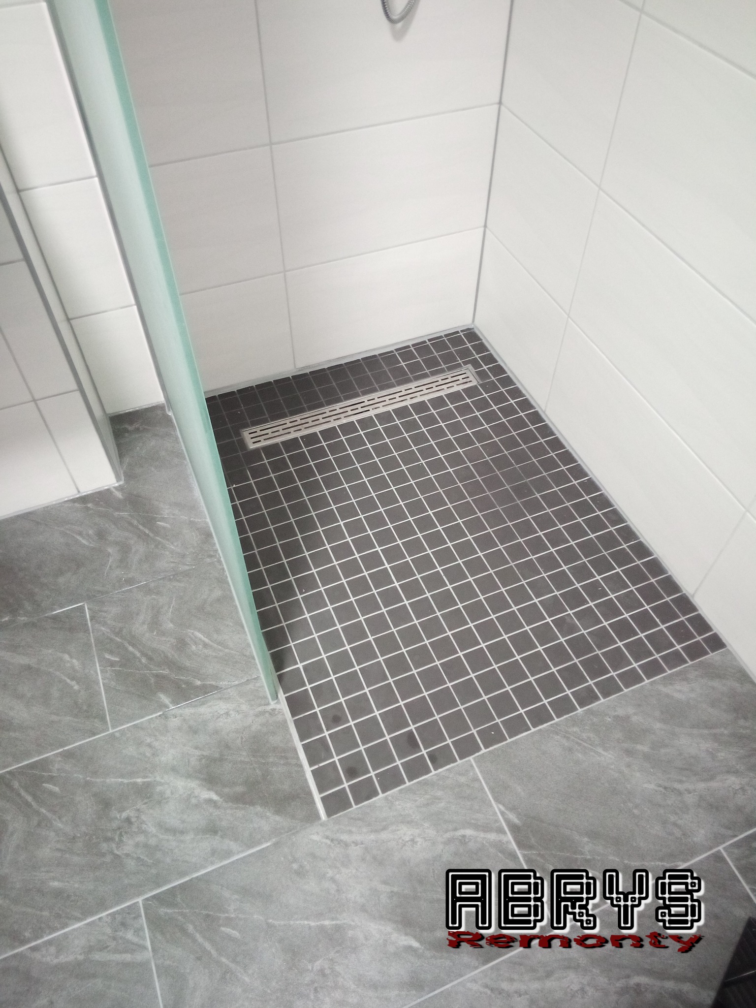 Nowoczesna kabina prysznicowa z szarymi płytkami mozaikowymi na podłodze brodzika i dużymi białymi płytkami na ścianach, oddzielona szklaną ścianką, z widocznym odpływem liniowym i szarymi płytkami...