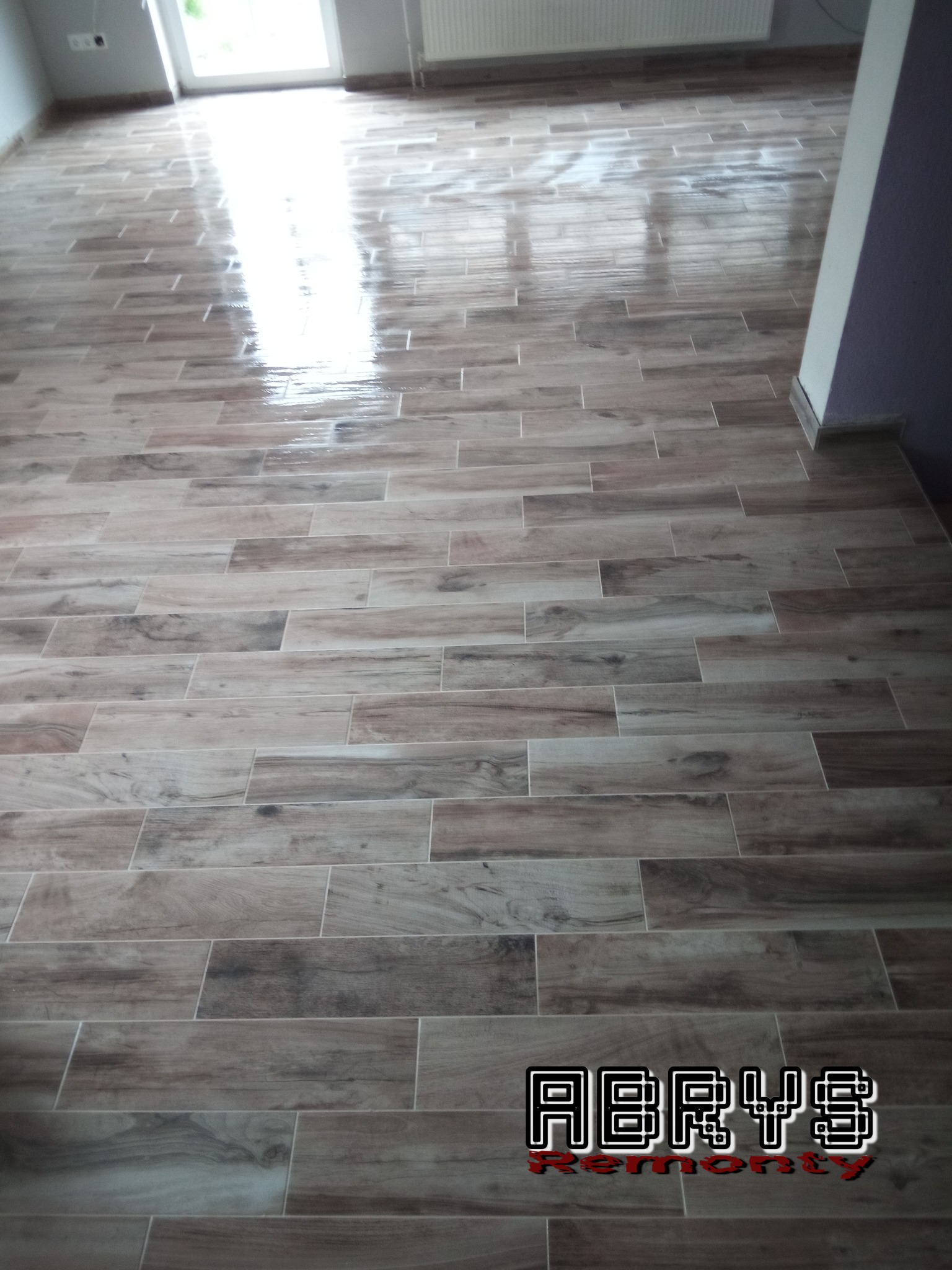 Nowo położona, błyszcząca podłoga z płytek imitujących drewno w jasnym pomieszczeniu z grzejnikiem i oknem, widoczne logo firmy remontowej w rogu.