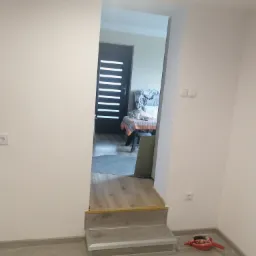 Wnętrze mieszkania po malowaniu ścian na biało, widoczne schodki z szarą okładziną, w tle fragment wyposażenia pokoju i czarne drzwi wejściowe.