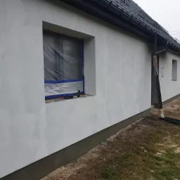 Świeżo otynkowana elewacja domu jednorodzinnego z zabezpieczonym oknem folią i taśmą malarską, widoczny fragment dachu z czarną dachówką i rynną.