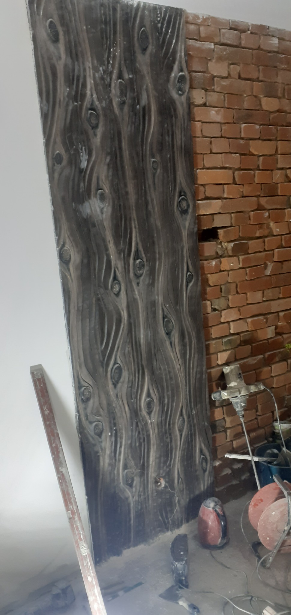 Dekoracyjna ściana z efektem drewna w szarym odcieniu, sąsiadująca z surową ścianą z czerwonej cegły, widoczne narzędzia budowlane i nieporządek na podłodze, sugerujące trwające prace wykończeniowe.