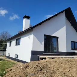 Biały dom z czarnym dachem i oknami, świeżo po remoncie elewacji, z widocznym rusztowaniem i stertą ziemi w ogródku.