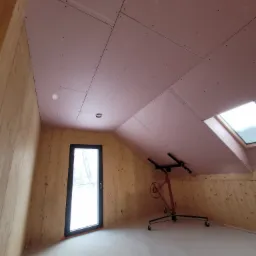 Montaż płyt kartonowo-gipsowych na skośnym suficie poddasza w Szczepanowie, widoczny podnośnik do płyt i drewniane wykończenie ścian.