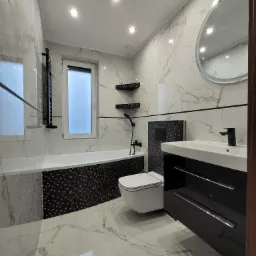 Elegancka łazienka w bieli i czerni z marmurowymi płytkami, okrągłym lustrem, podwieszaną toaletą i czarną armaturą.