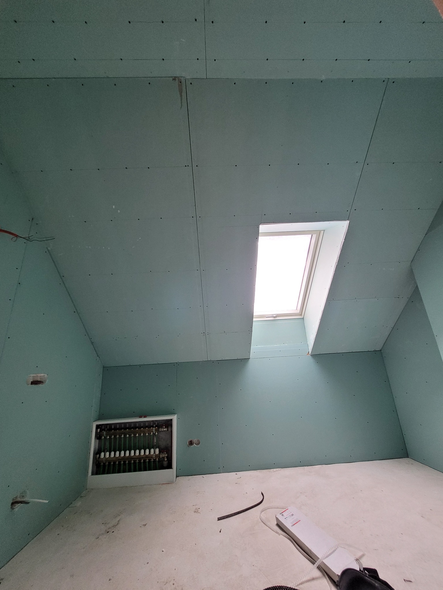 Wnętrze pokoju ze skośnym sufitem, wykończone zielonymi płytami gipsowo-kartonowymi, widoczne okno dachowe i rozdzielacz instalacji grzewczej w ścianie, surowa podłoga.