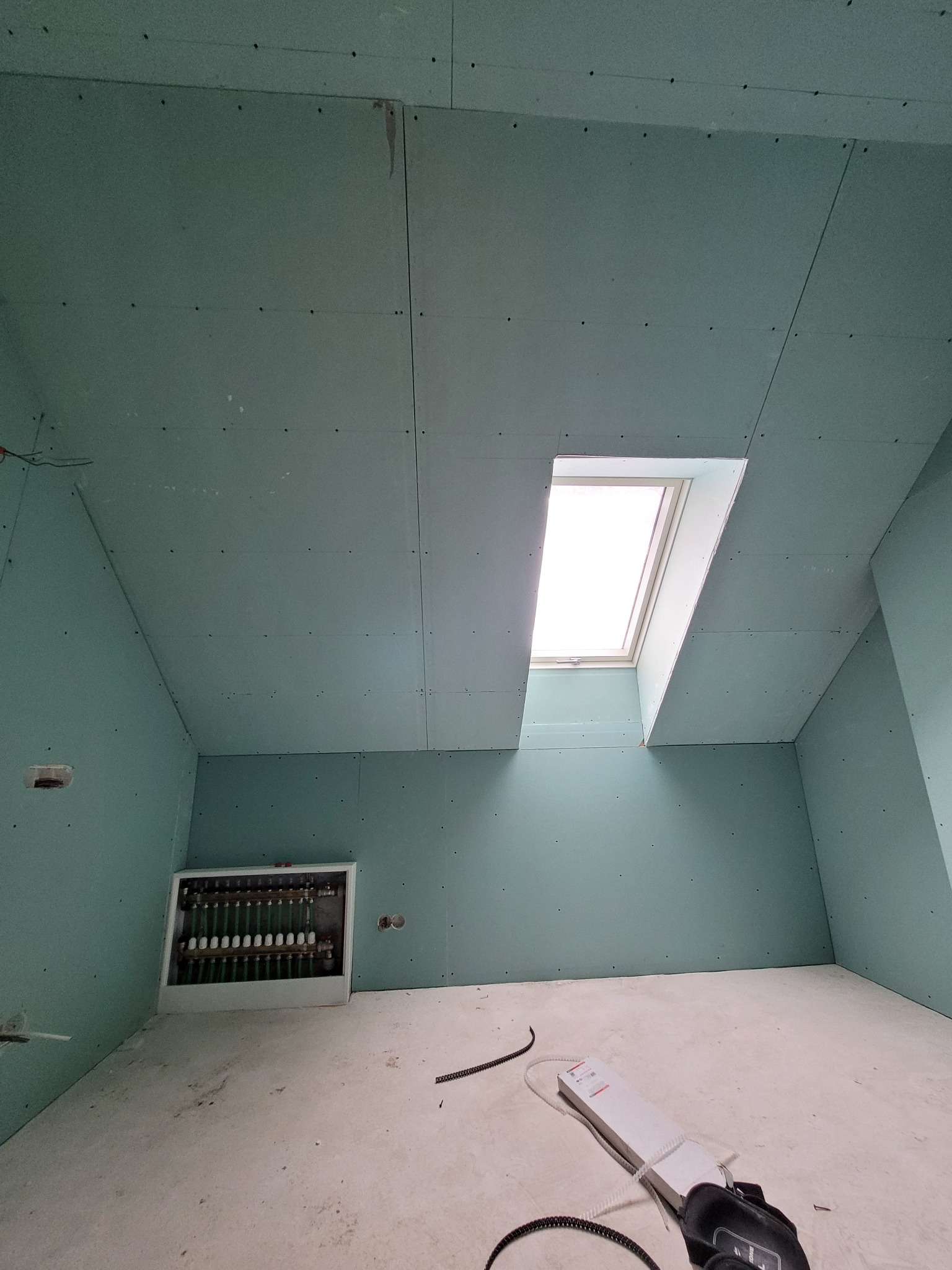 Poddasze z oknem dachowym w trakcie wykańczania płytami kartonowo-gipsowymi w kolorze zielonym, widoczna instalacja grzewcza w ścianie oraz narzędzia na podłodze.