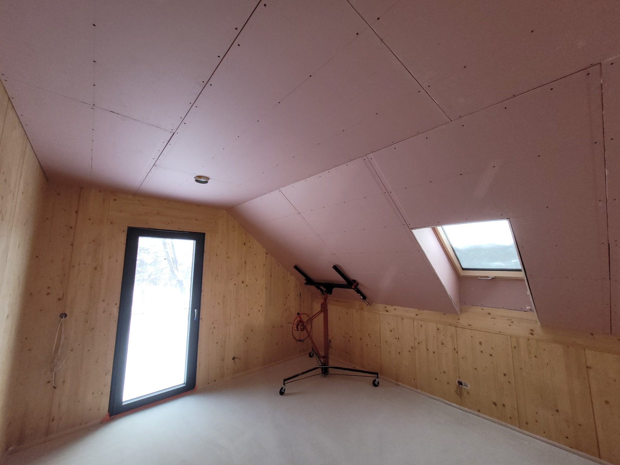 Poddasze z drewnianą boazerią i zamontowanymi płytami kartonowo-gipsowymi na suficie, widoczne okno dachowe i podnośnik do płyt. Płyty koloru różowego. Na ścianie widoczne wyprowadzone kable.