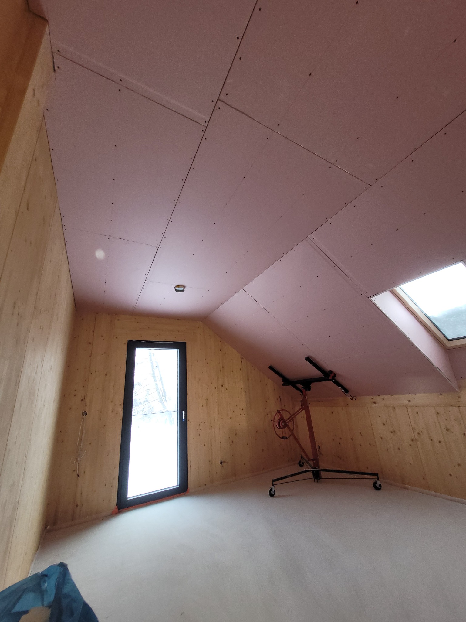 Montaż płyt kartonowo-gipsowych na skośnym suficie poddasza w Szczepanowie, widoczny podnośnik do płyt i drewniane wykończenie ścian.