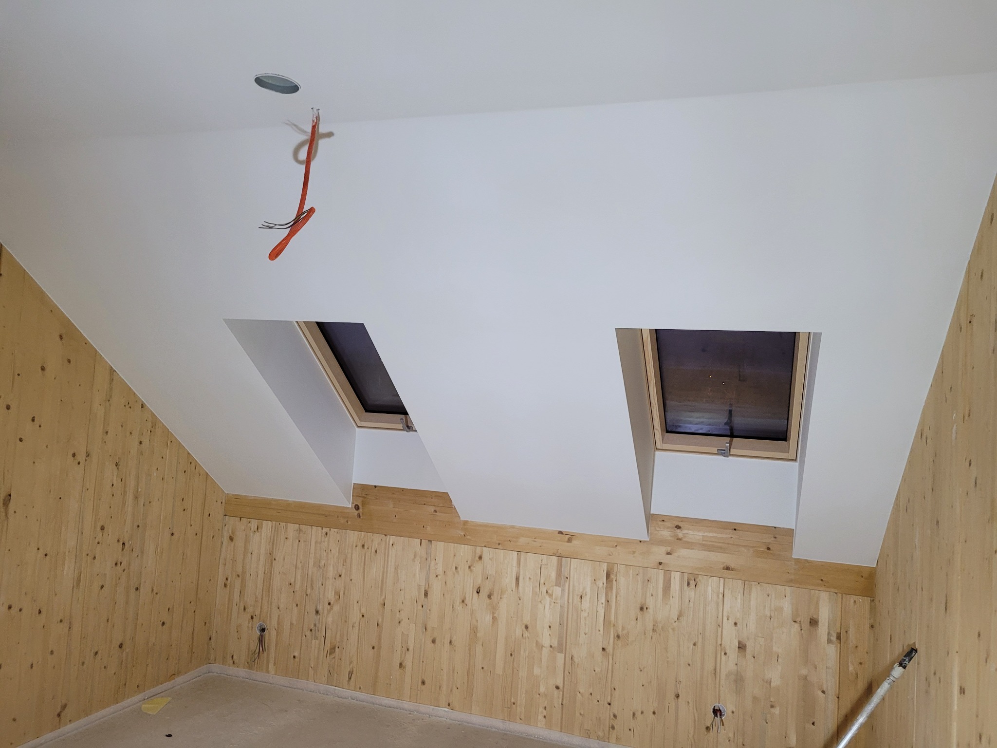 Poddasze z drewnianą boazerią, dwoma oknami dachowymi i białym sufitem z wystającym przewodem elektrycznym i otworem na oświetlenie