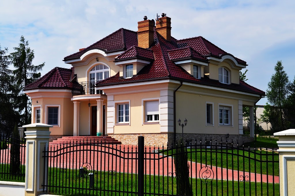 Elegancki, dwukondygnacyjny dom z beżową elewacją, czerwoną dachówką i czarnym, kutym ogrodzeniem, sfotografowany w słoneczny dzień.