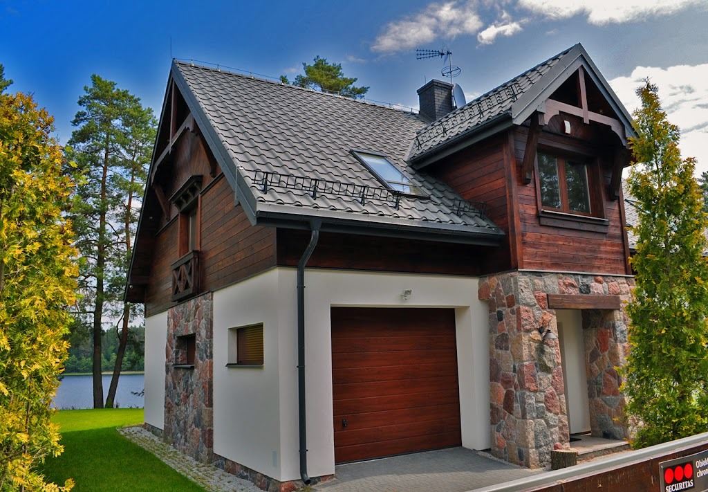 Dwukondygnacyjny dom z brązowego drewna z garażem i kamienną elewacją, grafitowy dach z oknem połaciowym, na tle zieleni i jeziora.