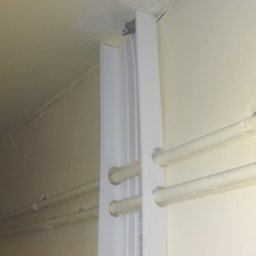 TELE Martin Jacek Martin - Biała listwa przypodłogowa z otworami na rury centralnego ogrzewania, zamontowana przy ścianie i suficie w pomieszczeniu o kremowych ścianach.