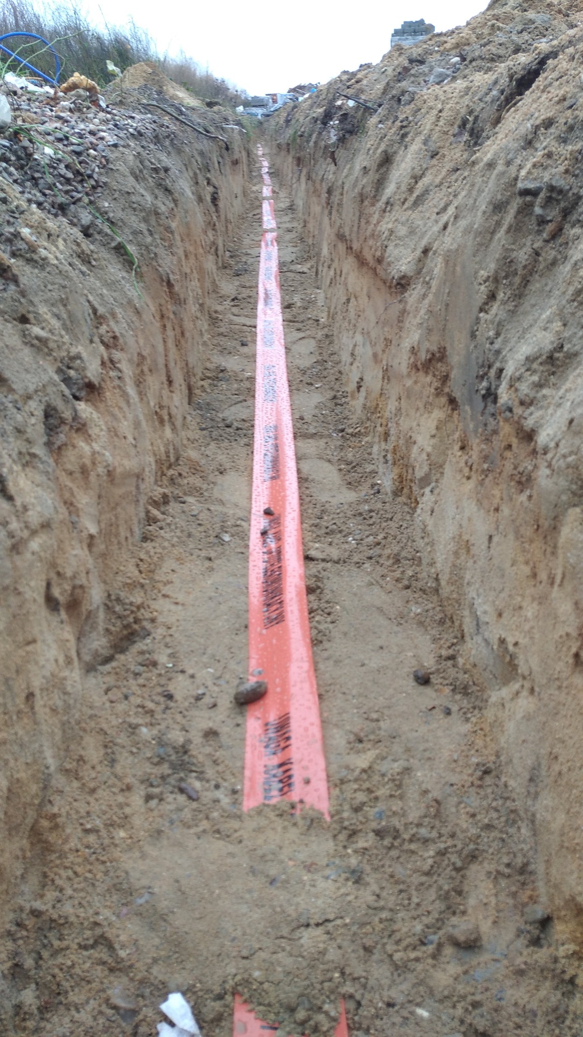 Wykop w ziemi z pomarańczową taśmą ostrzegawczą z napisem 'UWAGA KABEL' ułożoną wzdłuż wykopu, widok z góry.