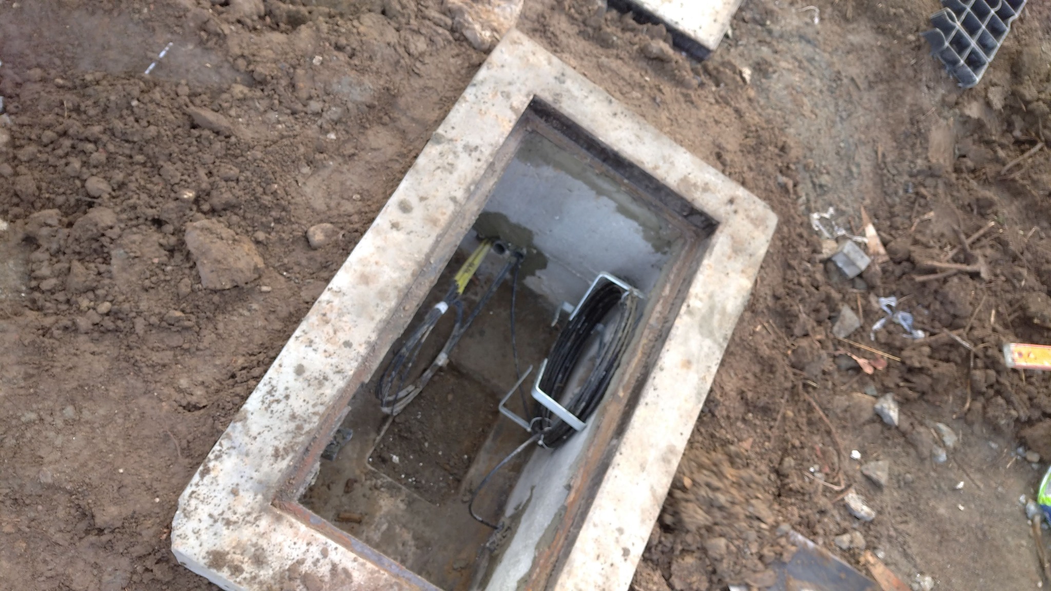 Betonowa studzienka rewizyjna z kablami telekomunikacyjnymi w ziemi, widok z góry, ziemia wokół wykopu, ułożony kabel wewnątrz studzienki.