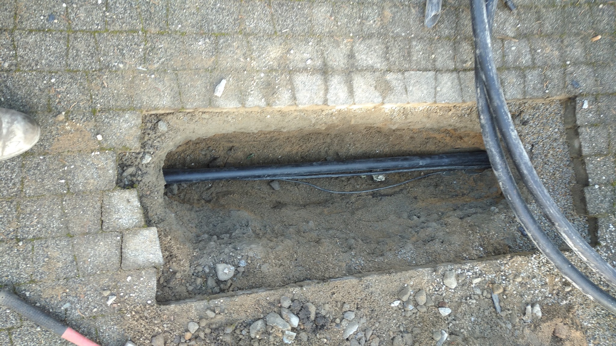 Wykop w chodniku z widocznymi czarnymi rurami w ziemi, obok leżące kable i fragment buta oraz młotek.