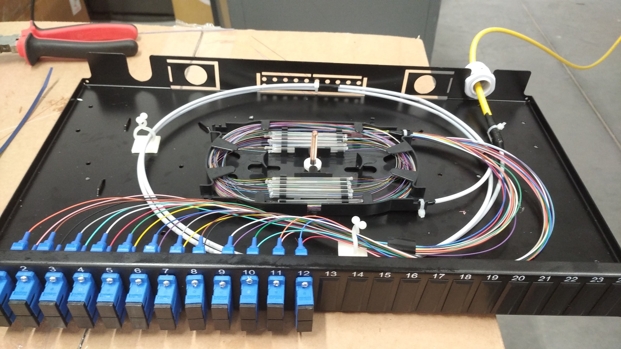 Panel krosowniczy światłowodowy z ponumerowanymi portami, kolorowymi kablami i spiralnie ułożonymi włóknami w czarnej obudowie.