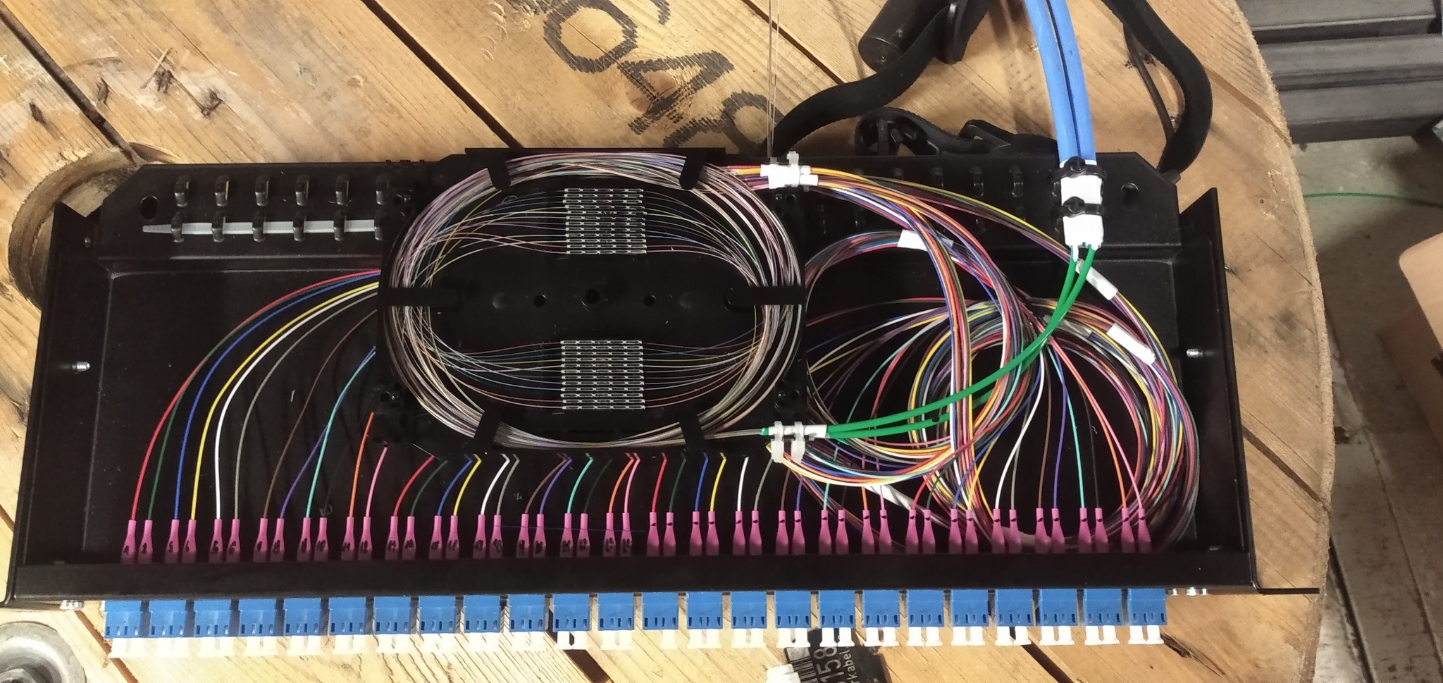 Panel krosowniczy światłowodowy z uporządkowanymi kablami w różnych kolorach, zakończonymi złączami, umieszczony na drewnianej powierzchni, ukazujący etap łączenia i organizowania przewodów...