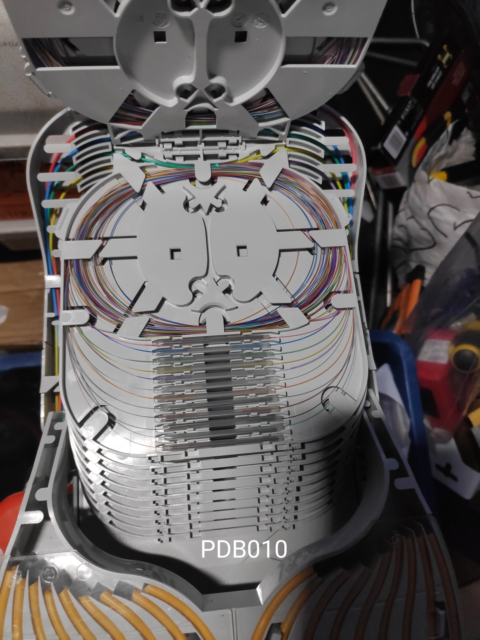 Otwarta skrzynka telekomunikacyjna z uporządkowanymi, kolorowymi kablami światłowodowymi, ułożonymi warstwowo, widoczne elementy mocujące i prowadnice kabli.