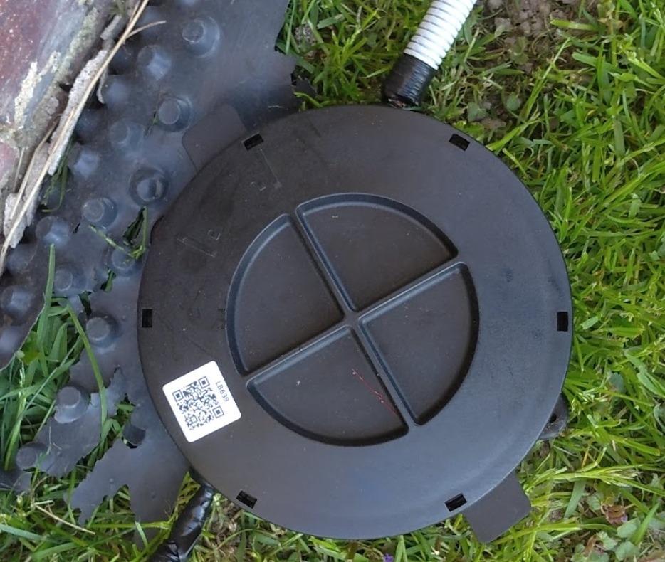 Czarna, okrągła puszka instalacyjna z naklejką QR code leży na trawie obok fragmentu karbowanej rury ochronnej i elementu drenażowego.