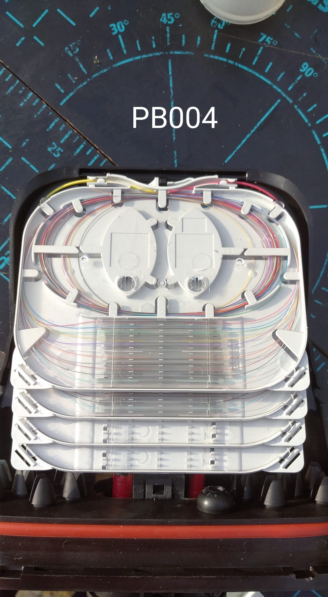 Rozdzielnica światłowodowa PB004 z widocznymi włóknami w różnych kolorach ułożonymi wewnątrz, umieszczona na tle z podziałką kątową.