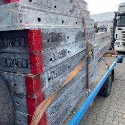 Nie Podano - Metalowe szalunki budowlane o wymiarach 30x150, 50x150 i 75x150, zabezpieczone pasami transportowymi na niebieskiej przyczepie. Widoczny fragment opony przyczepy i kostka brukowa podłoża.