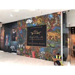 Nie Podano - Kolorowa grafika ścienna z logo 'NAHLA COFFEE & CHOCOLATE 100% COSTA RICAN' w przestrzeni handlowej, obok witryny sklepowej i siedzącego pracownika w kamizelce odblaskowej.