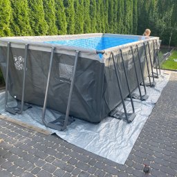 Nie Podano - Rozkładany basen ogrodowy Intex z szarą obudową i niebieskim wnętrzem, rozłożony na srebrnej plandece na kostce brukowej, z rzędem wysokich tui w tle i osobą w środku.