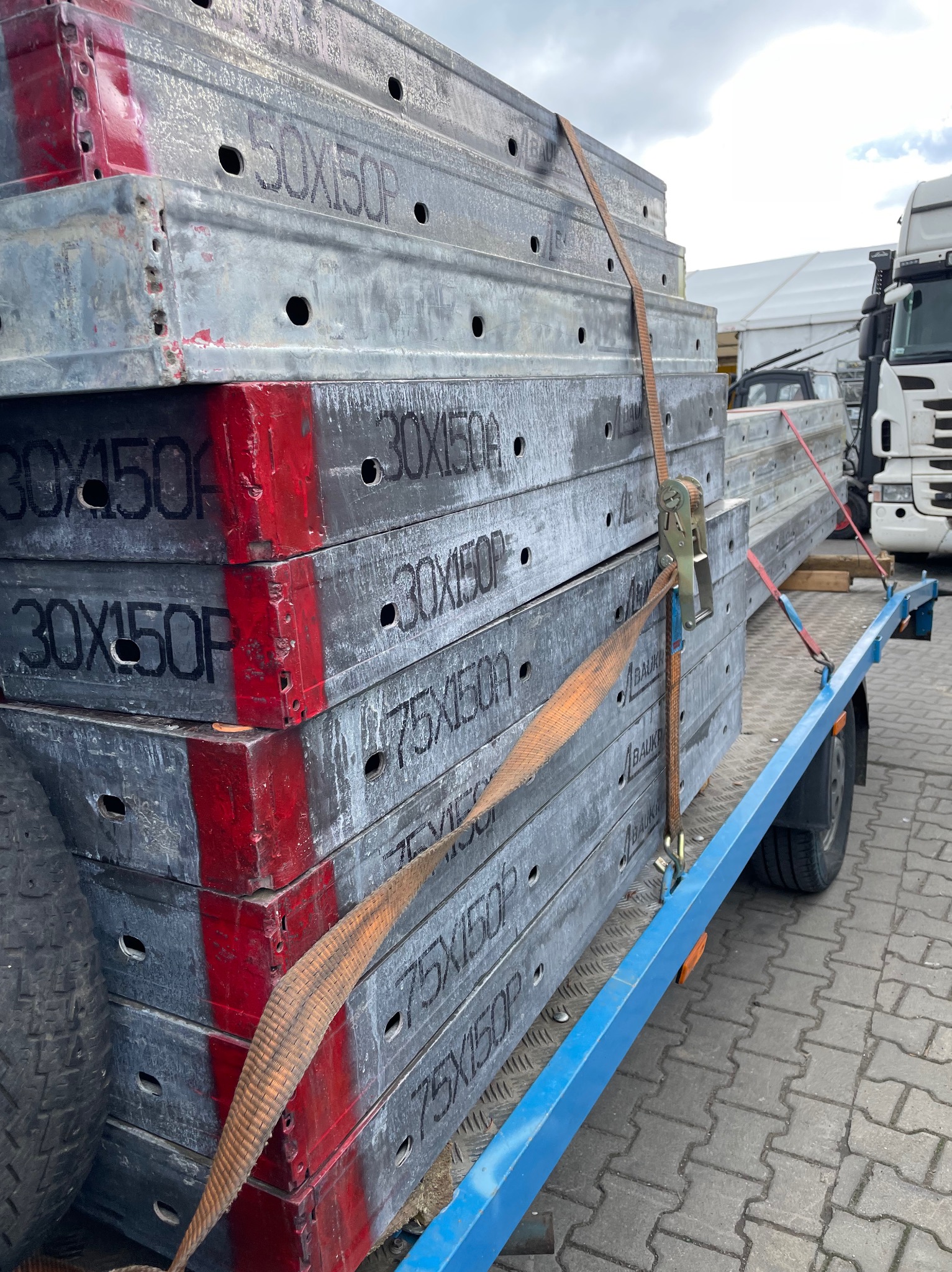 Metalowe szalunki budowlane o wymiarach 30x150, 50x150 i 75x150, zabezpieczone pasami transportowymi na niebieskiej przyczepie. Widoczny fragment opony przyczepy i kostka brukowa podłoża.
