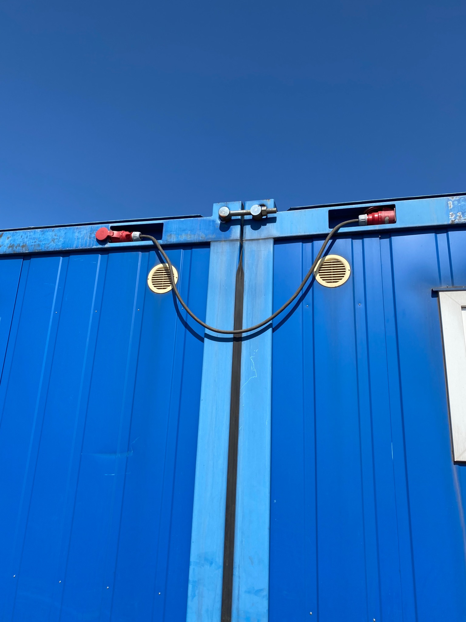 Fragment niebieskiego kontenera z widocznymi gniazdami elektrycznymi, wentylacją i elementami mocującymi na tle błękitnego nieba.