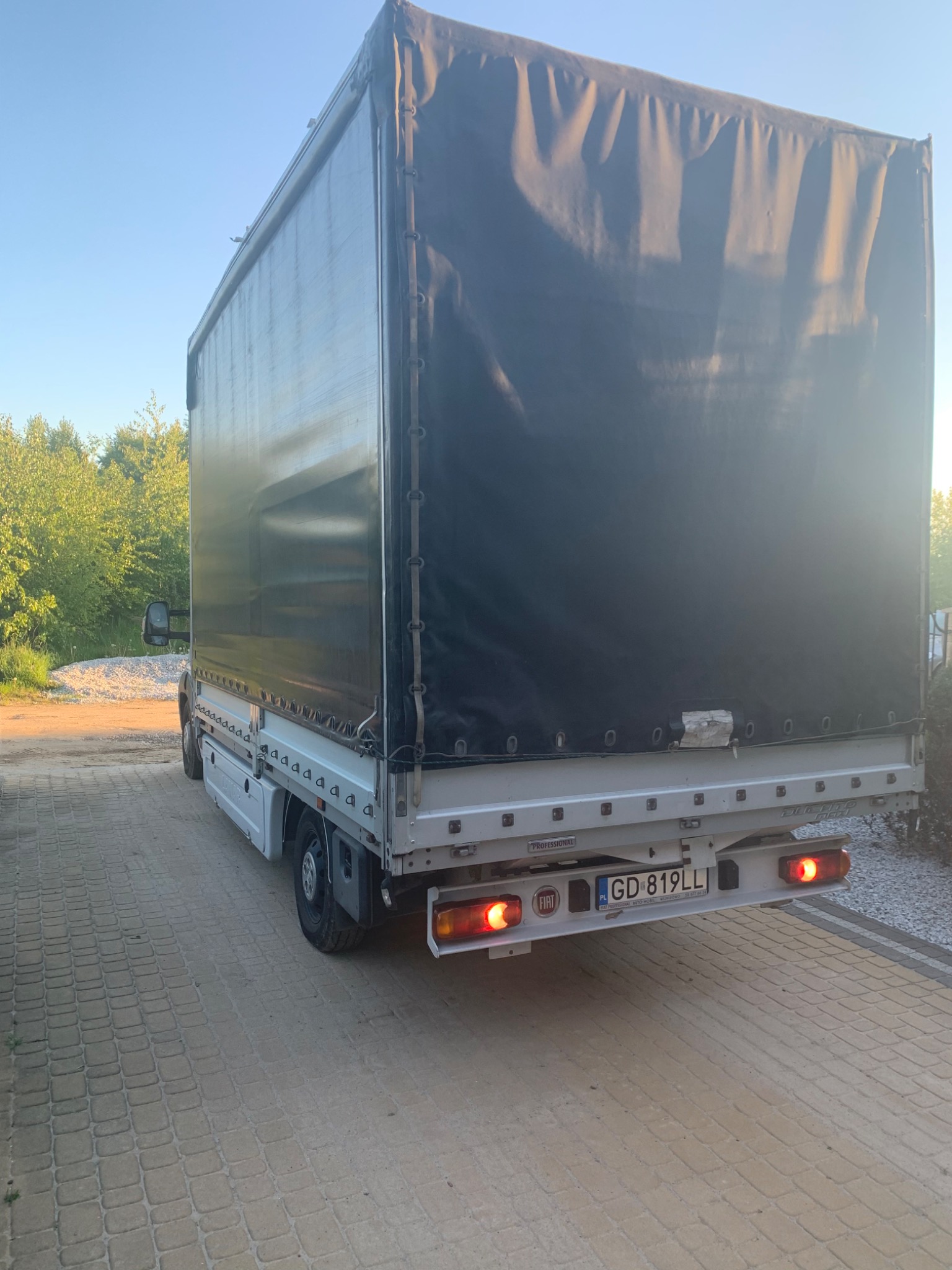 Tył białego busa z czarną plandeką, zaparkowanego na brukowanej drodze, widoczne światła stopu i tablica rejestracyjna GD 819LL, w tle zieleń drzew.