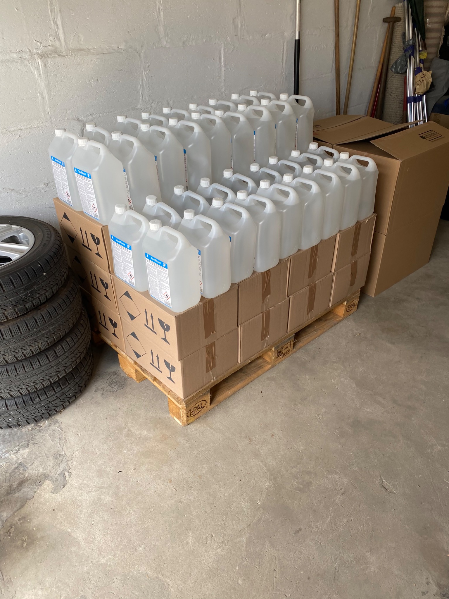 Paleta z kartonami, na których ułożone są plastikowe kanistry z płynem, obok opony samochodowe; widoczne symbole ostrzegawcze na kartonach.
